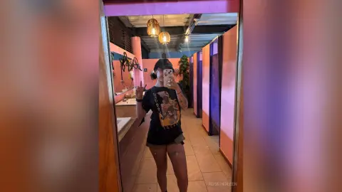 RossiNoleen takes a mirror selfie wearing a black graphic t-shirt and black shorts while standing in a room with orange walls.