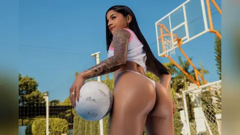 KendallRosy poses outdoors with a white soccer ball, wearing a white crop top and white thong that shows off her bare, tanned buttocks and tattooed arms.