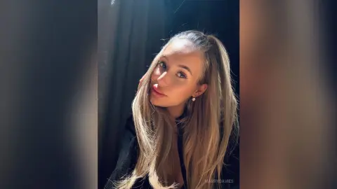 MarryDavies poses with her hand against her head, showing her long blonde hair and face in soft lighting.