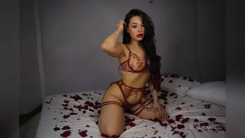 Young woman in red lace lingerie sitting on a bed covered in rose petals in a moody bedroom setting.