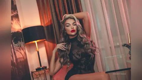 Anastasia poses in a black lace bodysuit with long, wavy blonde hair and bright red lipstick.