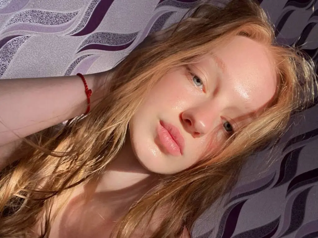 Close-up portrait of a blonde woman with glowing skin lying on a patterned purple fabric in warm sunlight.