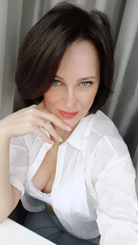 Smiling woman with short brown hair wearing a white button-down shirt in a bright indoor setting.
