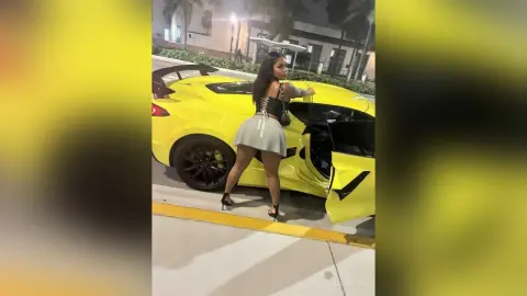 Stylish woman in a grey mini skirt and black top posing next to a bright yellow luxury sports car at night.