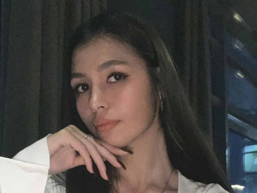 Young woman with long dark hair wearing a white blouse in a moody, dimly lit indoor setting.