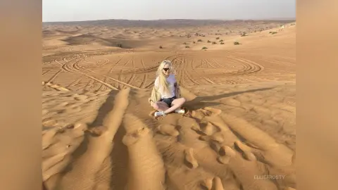 EllieFrosty sits on a sand dune in a white t-shirt and black shorts, surrounded by desert sand patterns.