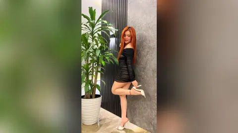Red-haired woman in a sleek black mini dress posing by a potted plant in a modern indoor setting.