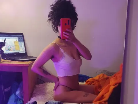 Hathorlove poses in a white lace camisole and black thong, showing her curly hair and tattoos while sitting on a white bed next to a laptop.