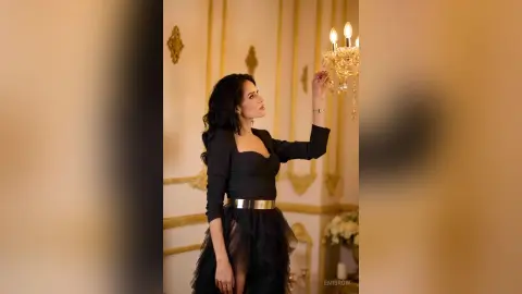 Elegant woman in a black long-sleeve dress with a gold belt posing by a crystal chandelier in a luxury room.