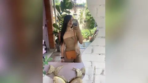 Young woman in a tan sleeveless jumpsuit taking a selfie in a bright, sunlit indoor garden setting.