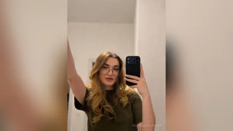 Young woman with long blonde hair wearing glasses and an olive green t-shirt taking a mirror selfie indoors.