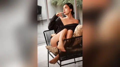 LucyMartin sits on a chair wearing a black off-the-shoulder top, posing with one hand near her lips and her bare legs crossed.