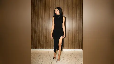 Elegant woman in a black sleeveless maxi dress posing against a textured wooden wall in a moody setting.