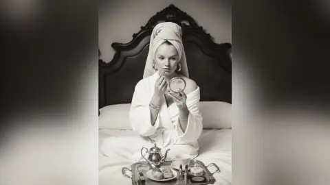 Elegant woman in a white bathrobe and hair towel applying makeup in a cozy bedroom setting.