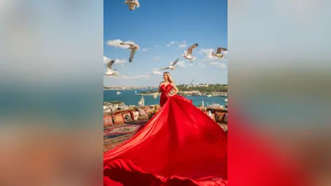 Anita poses in a long red dress on a rooftop overlooking the sea with white birds flying in the blue sky.