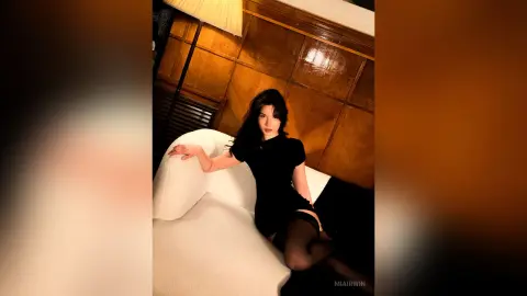 Elegant woman in a black dress relaxing on a white sofa in a warm, moody wood-paneled room.