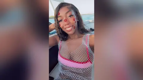 NatyGlow smiles wearing a white mesh tank top with pink trim, showing her cleavage while sitting outdoors near the water.