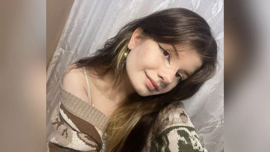 Young woman with winged eyeliner and a brown hair bow posing in a cozy indoor setting.