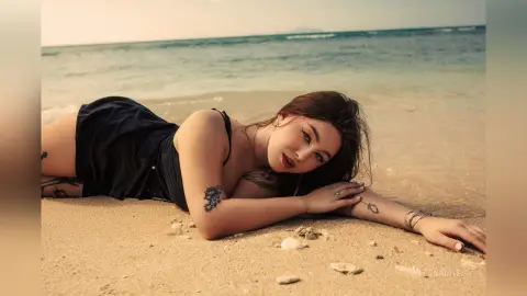 Tattooed woman in a black summer dress relaxing on a sunny beach with ocean waves in the background.