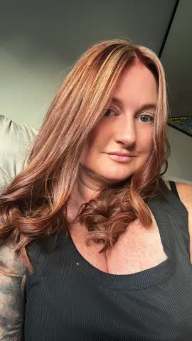 Smiling woman with long wavy reddish-brown hair wearing a black ribbed tank top in a cozy indoor setting.