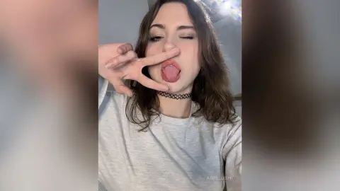 AriaLush sticks her tongue out while making a peace sign with her hand, wearing a grey t-shirt and a black lace choker.
