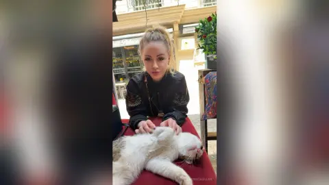 Blonde woman in a black jacket petting a fluffy white cat at an outdoor cafe terrace.