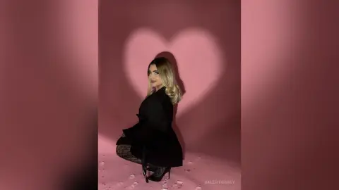 Stylish woman in a black blazer and patterned tights posing against a pink wall with heart-shaped shadow lighting.