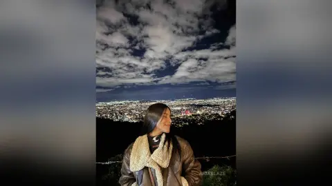 AriaBlaze smiles in a brown shearling jacket against a night city skyline and cloudy sky.