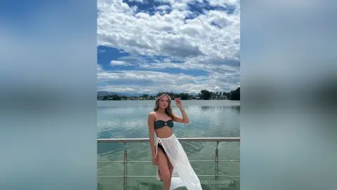 Young woman in a dark bikini and white sarong standing by a lake under a cloudy blue sky.