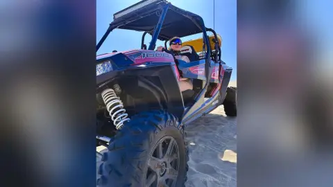 Adventurous woman in sunglasses driving a blue Polaris RZR off-road vehicle through desert sand dunes.