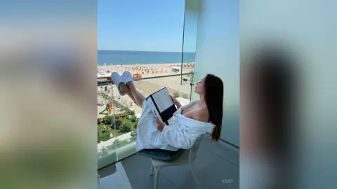 Relaxed woman in a white robe reading a book on a sunny balcony overlooking the ocean beach.