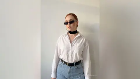 Redheaded woman in a white button-down shirt and black choker posing against a minimalist white wall.
