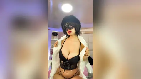 Glamorous woman in black lace lingerie and a masquerade mask wearing an open white shirt in a bright bedroom.