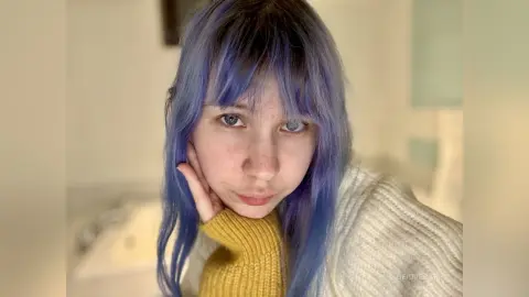Young woman with blue hair and a yellow sweater looking thoughtful in a bright indoor setting.
