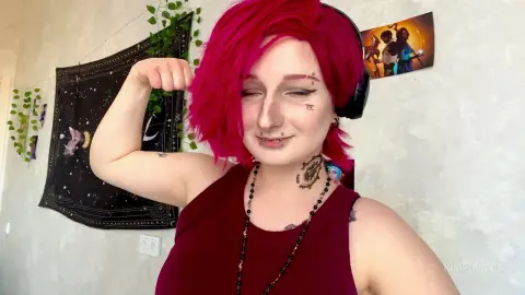 Smiling woman with pink hair and piercings wearing a maroon tank top in a cozy room.