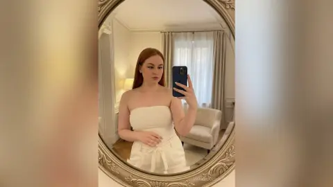 Redheaded woman in a white strapless dress taking a mirror selfie in a bright, modern living room.