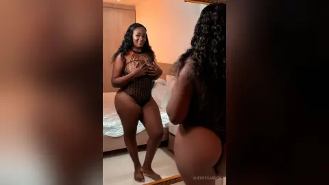 Curvy woman in a black sheer bodysuit posing in a bedroom mirror for a confident lifestyle portrait.
