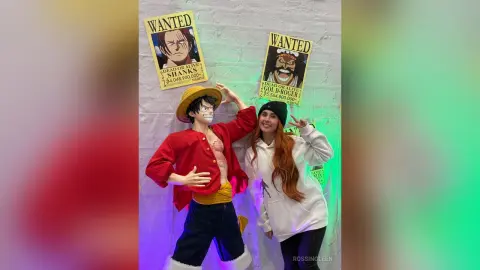 Smiling fan in a white hoodie posing next to a life-size Monkey D. Luffy cosplay and One Piece wanted posters.