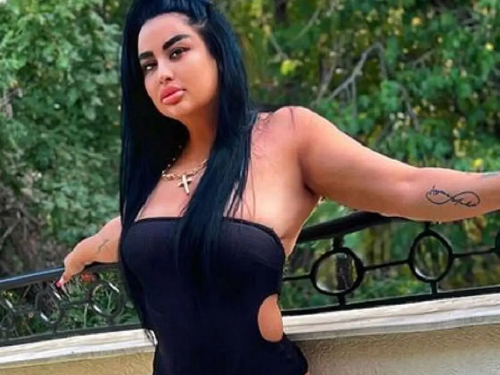 Curvy woman in a black strapless bodysuit posing outdoors against a lush green garden background.