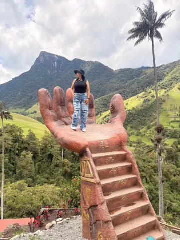 Victoria_Velvet stands on a large red hand statue overlooking a lush green mountain valley with palm trees.