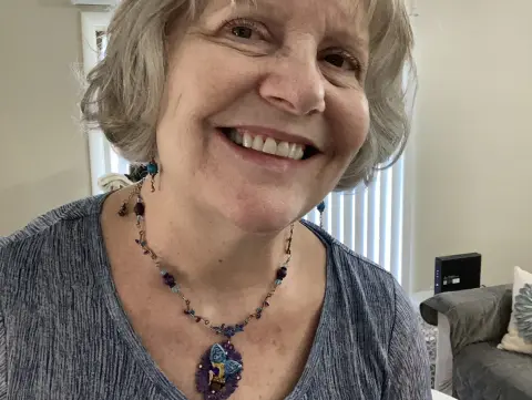 Smiling mature woman with short grey hair wearing a blue butterfly pendant necklace in a bright indoor setting.