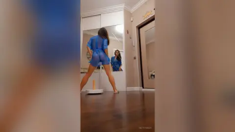 Young woman in a blue sheer outfit posing in a modern bedroom mirror reflection.