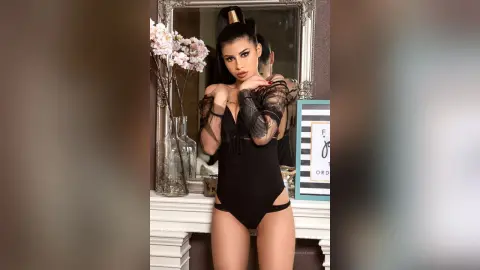 Glamorous woman in a black sheer bodysuit posing by a mirror with pink flowers in a chic indoor setting.