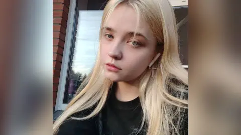 Blonde woman with cross earrings wearing a black t-shirt posing outdoors in natural light.