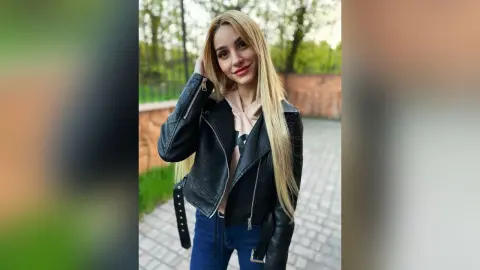 Smiling blonde woman in a black leather jacket and beige top posing outdoors in a park setting.