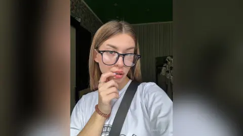 Young woman with glasses and white t-shirt eating a lollipop in a cozy indoor setting.