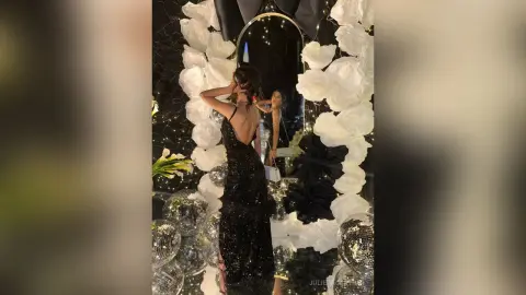 Elegant woman in a black sequin evening gown posing by a floral mirror display at a glamorous gala event.