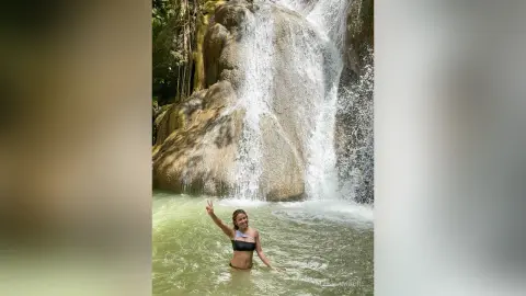 Smiling woman in a black bikini making a peace sign in a tropical jungle waterfall pool.