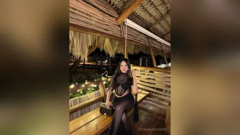Elegant woman in a black sheer cocktail dress posing in a rustic tropical bamboo hut at night.