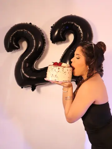 Young woman in a black dress celebrating her 22nd birthday with a cake in front of large black number balloons.